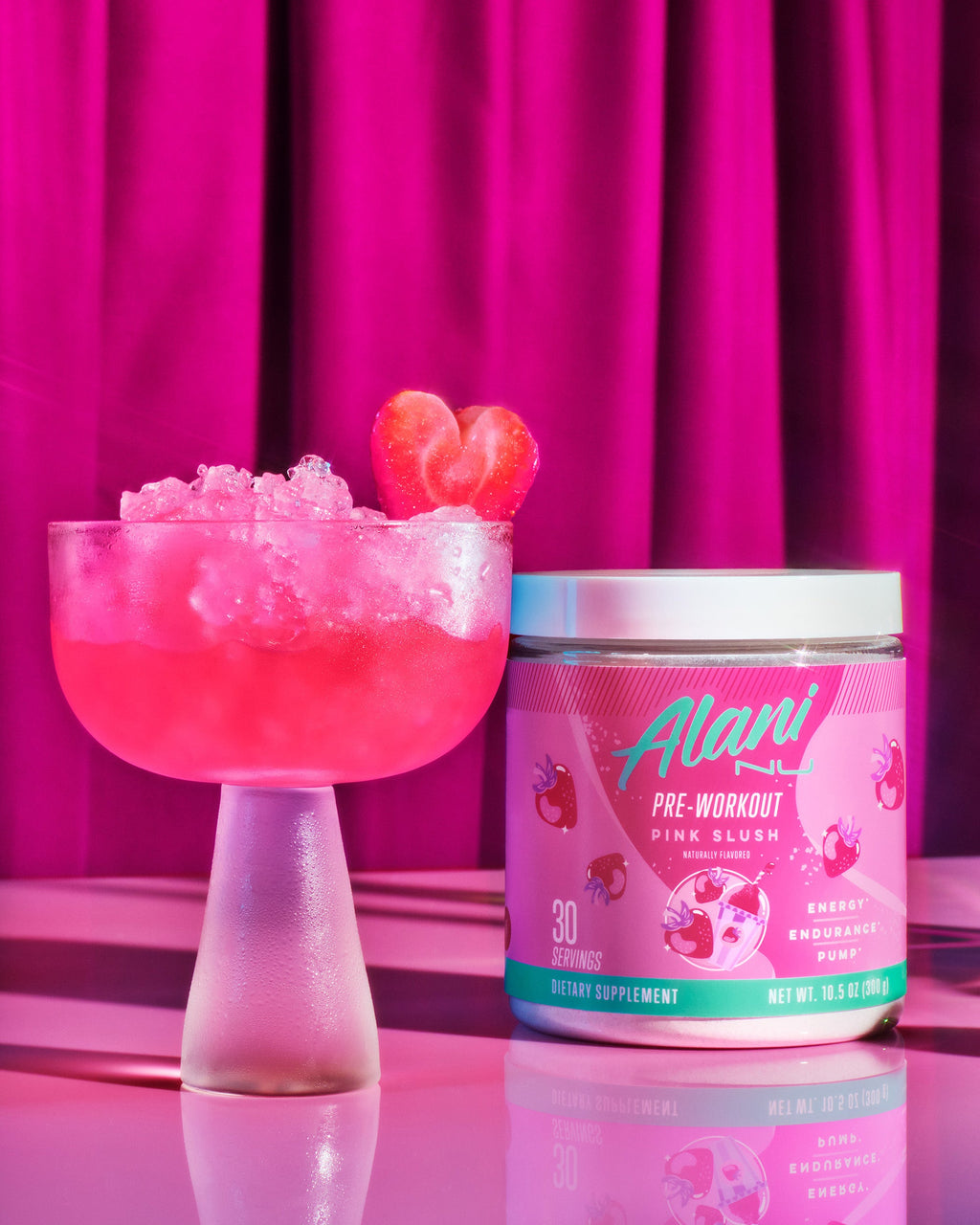 A tub of Alani Nu Pink Slush Pre-Workout next to a frosted coup glass filled with pebble ice and pink liquid garnished with a heart-shaped strawberry.